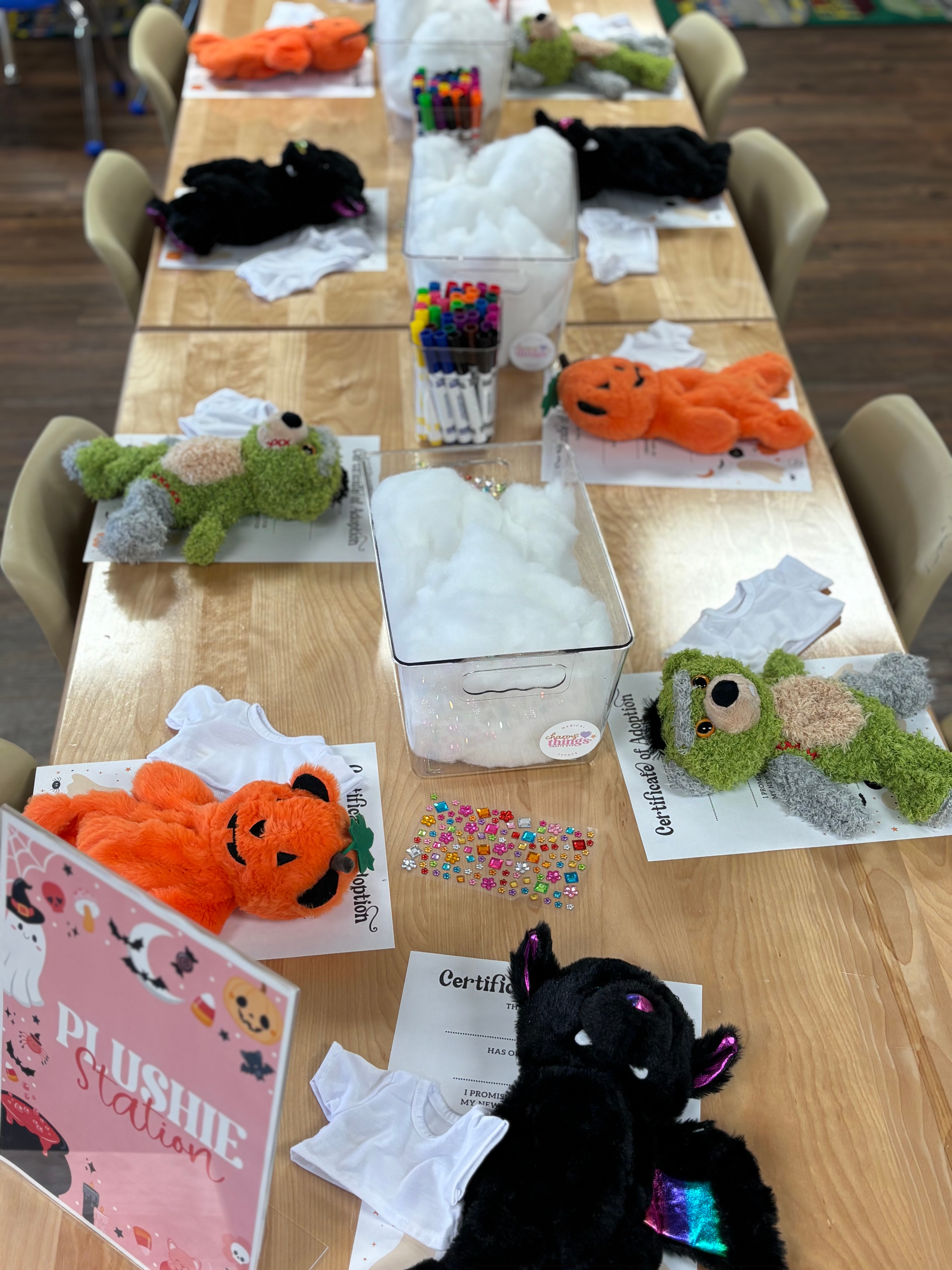 Table with plush toys, tissues, and craft supplies, including colorful markers and paper, in a room with chairs around the table.