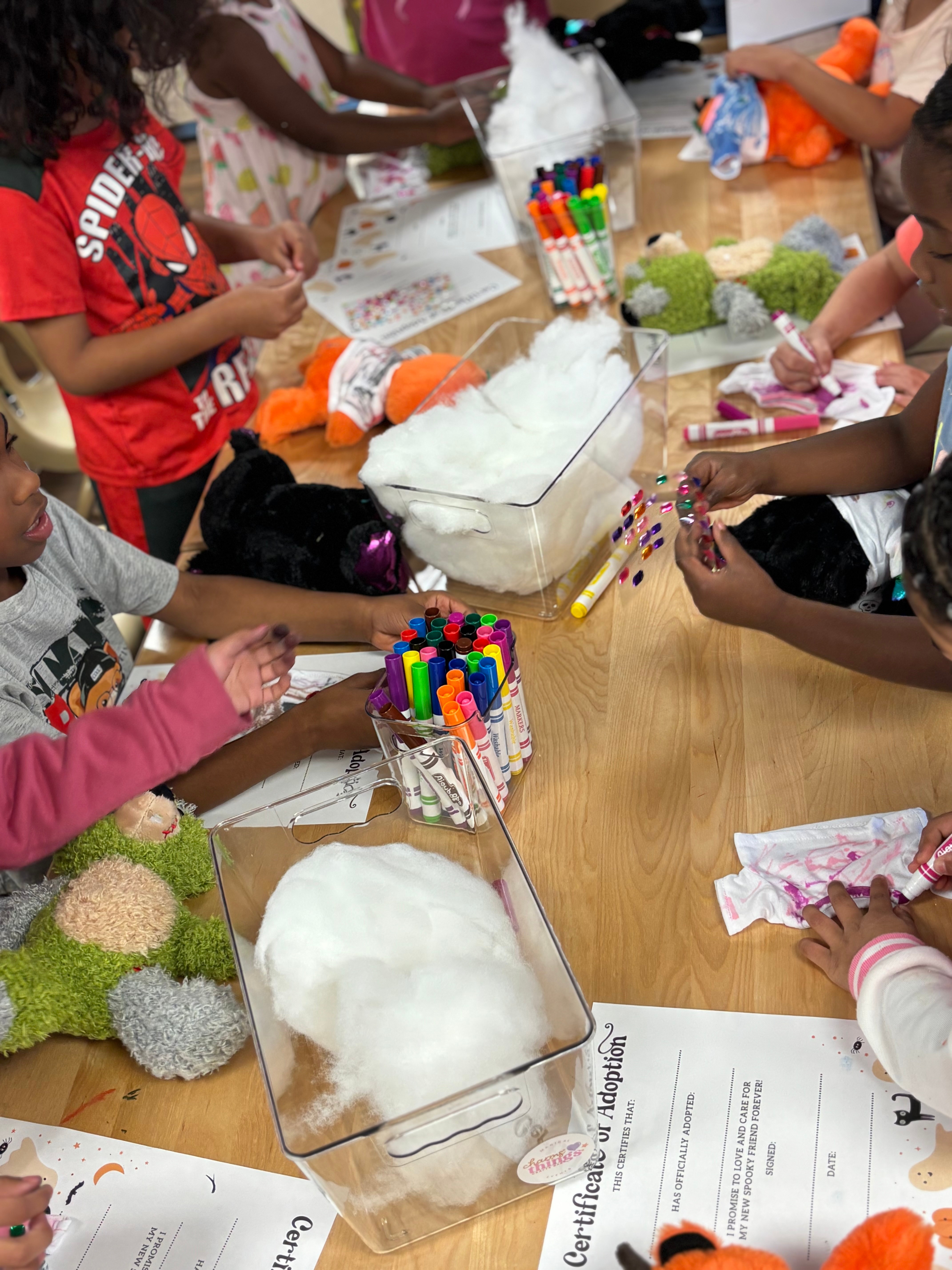 Children gather around a table with craft supplies, including markers, paper, and fluffy materials, engaged in a creative activity.