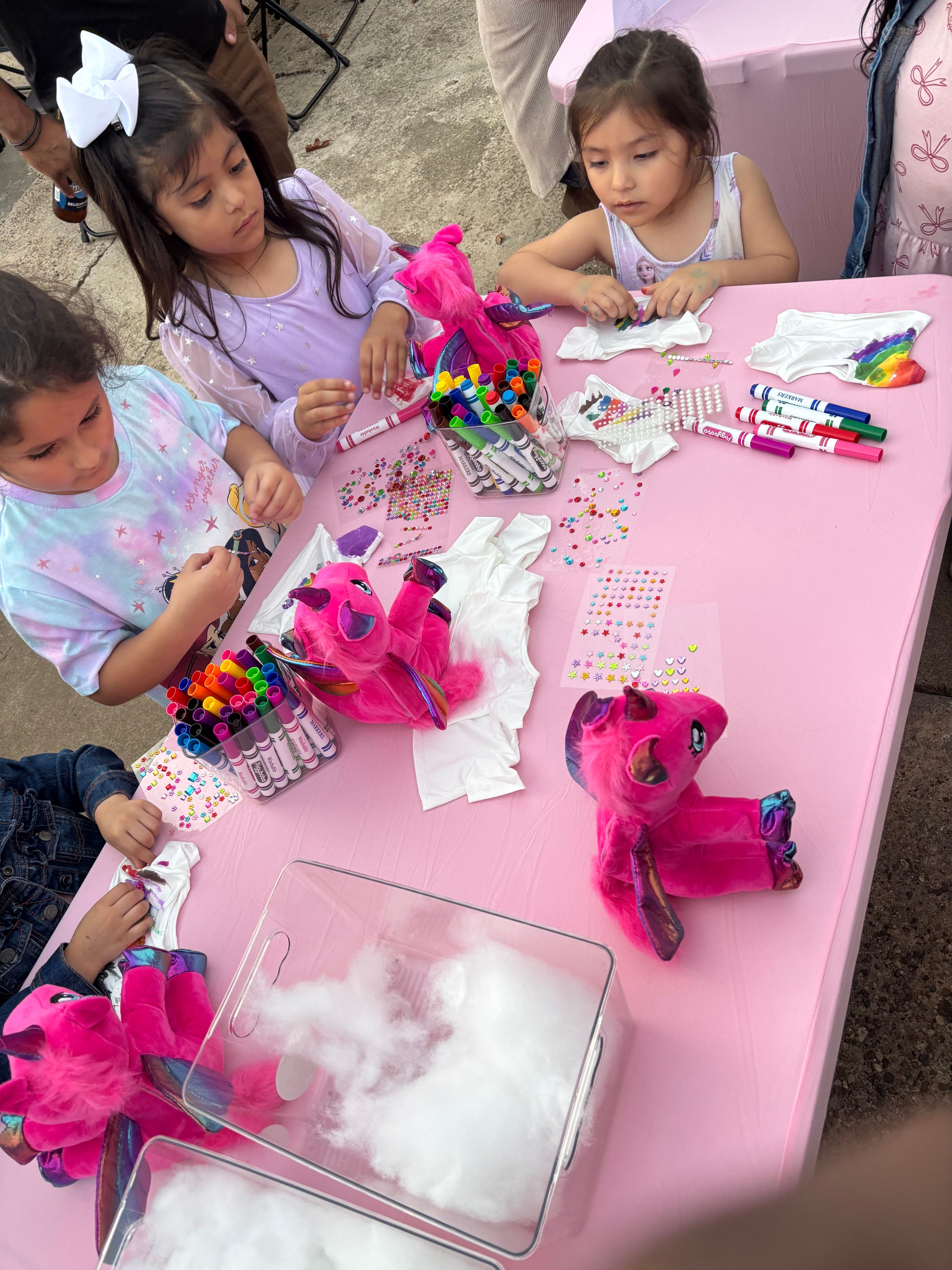 Two children at a pink table with colorful markers, plush toys, and paper crafts, smiling and engaging in activities.