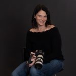 Woman sitting with a camera, smiling, wearing a black top and jeans, against a dark background.