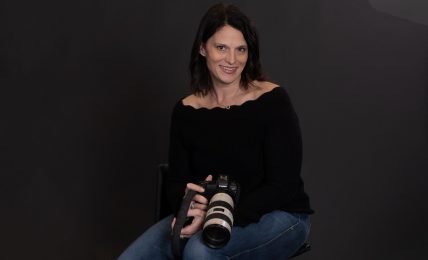 Woman sitting with a camera, smiling, wearing a black top and jeans, against a dark background.