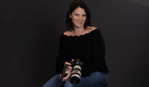 Woman sitting with a camera, smiling, wearing a black top and jeans, against a dark background.