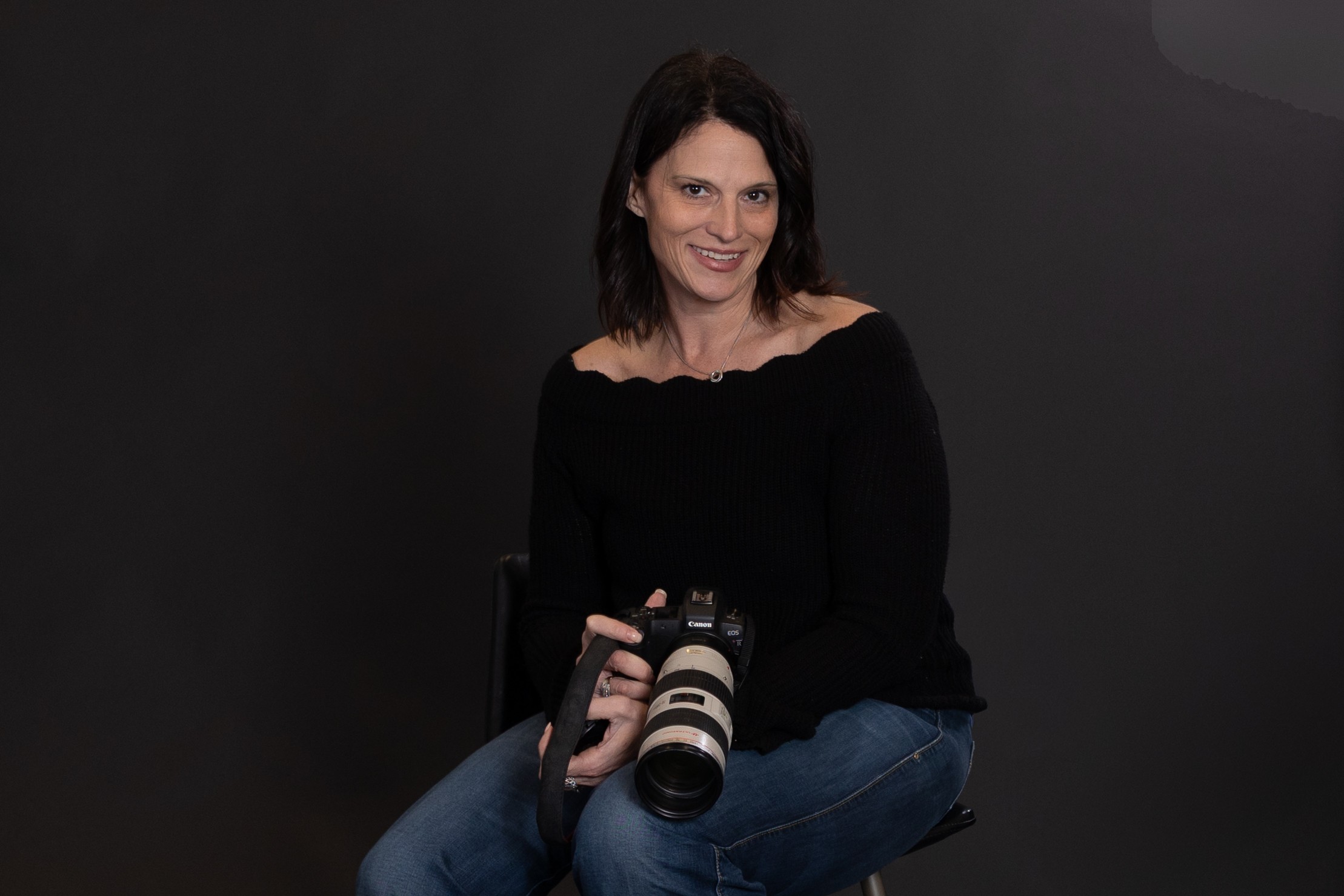 Woman sitting with a camera, smiling, wearing a black top and jeans, against a dark background.