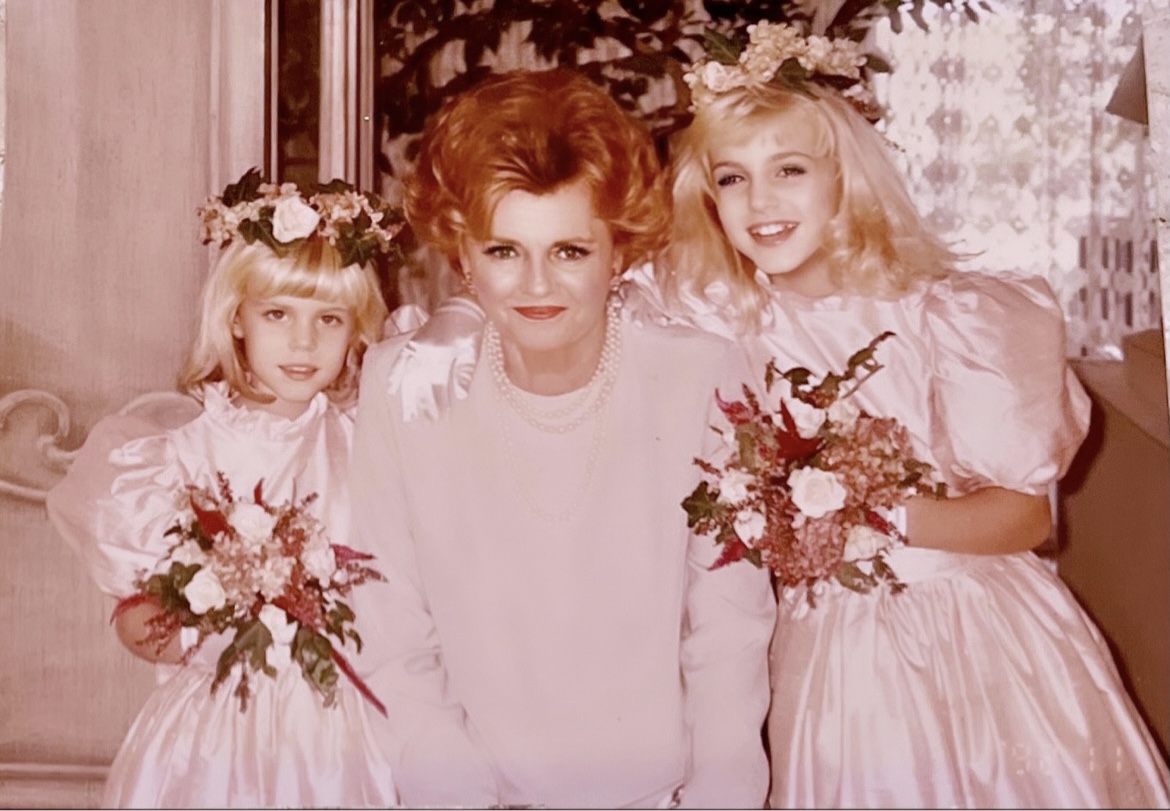 Three women in pink dresses holding bouquets, with a woman in the center, all smiling, indoors with floral decor