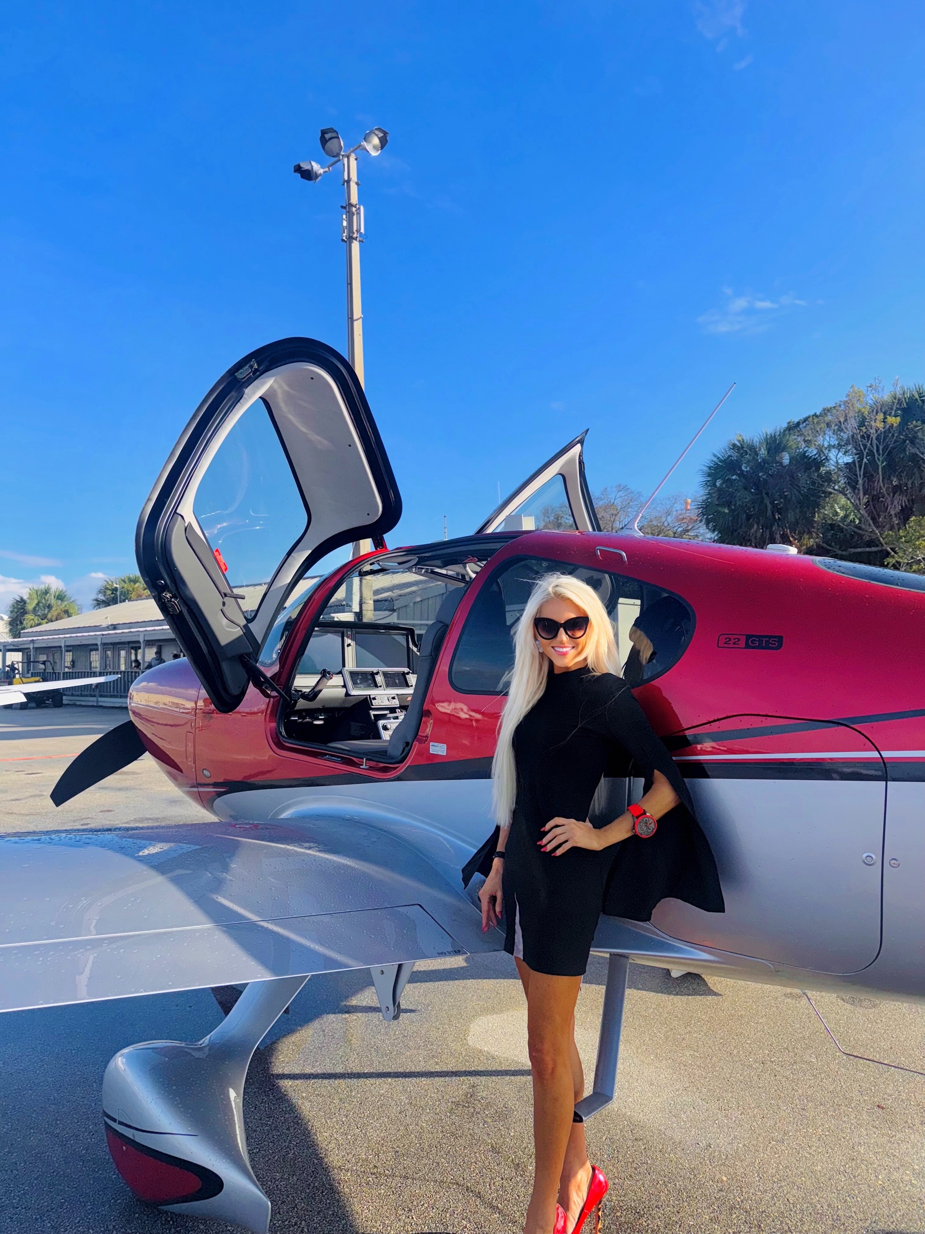 Woman with blonde hair and sunglasses standing next to a red and gray aircraft with open cockpit door, outdoors on sunny day.