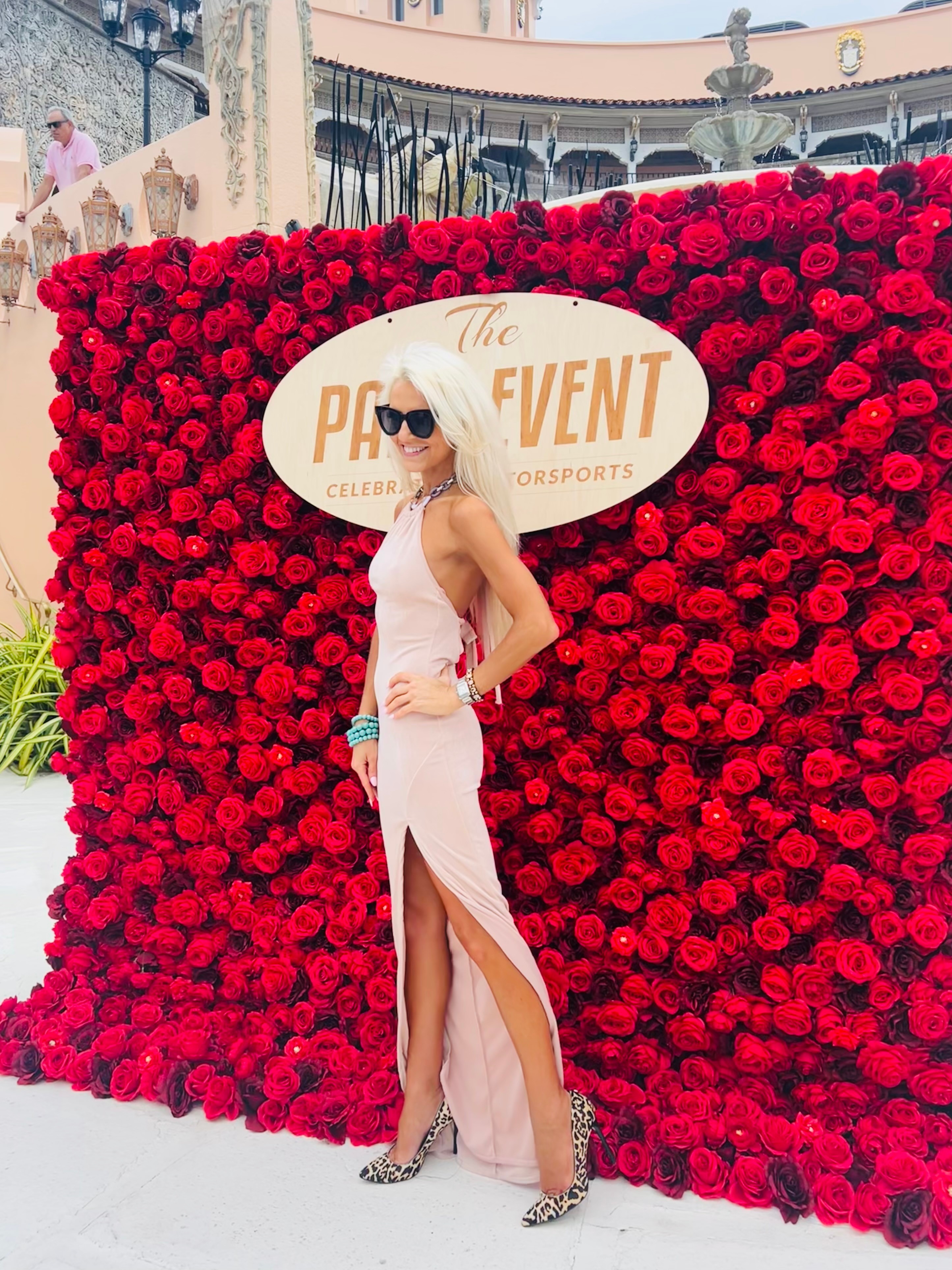 Woman in white dress with slit, sunglasses, and heels standing in front of a red flower wall with event sign.