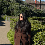 Woman with long dark hair wearing sunglasses and a brown coat standing outdoors near greenery, with a bridge in the background.