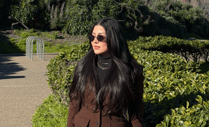 Woman with long dark hair wearing sunglasses and a brown coat standing outdoors near greenery, with a bridge in the background.