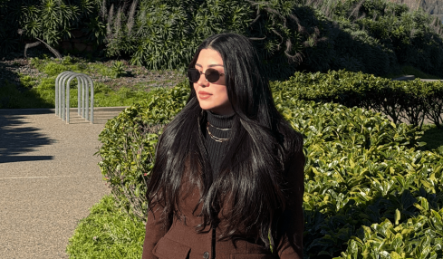 Woman with long dark hair wearing sunglasses and a brown coat standing outdoors near greenery, with a bridge in the background.