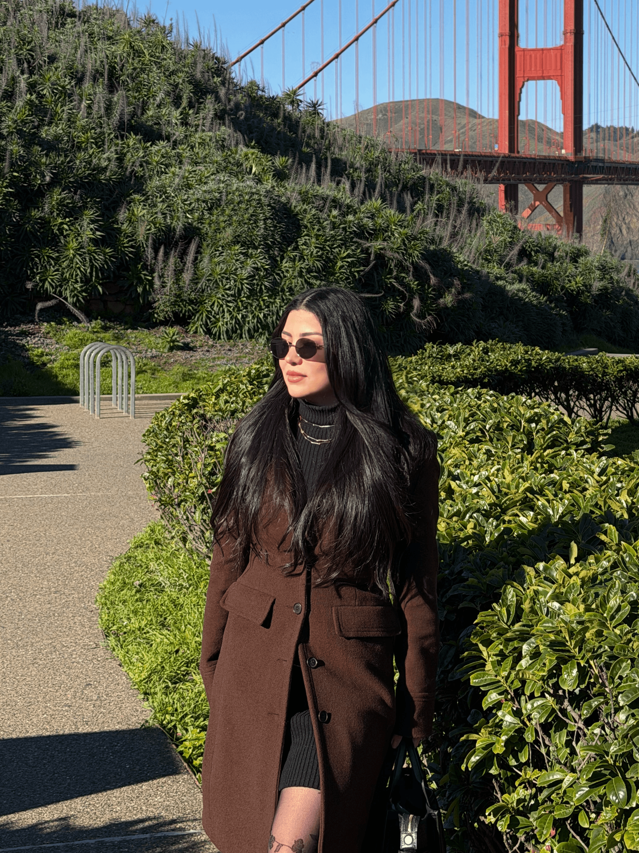 Woman with long dark hair wearing sunglasses and a brown coat standing outdoors near greenery, with a bridge in the background.