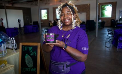 Smiling woman holding a small box in a decorated event space with tables and a chalkboard sign.