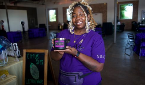 Smiling woman holding a small box in a decorated event space with tables and a chalkboard sign.