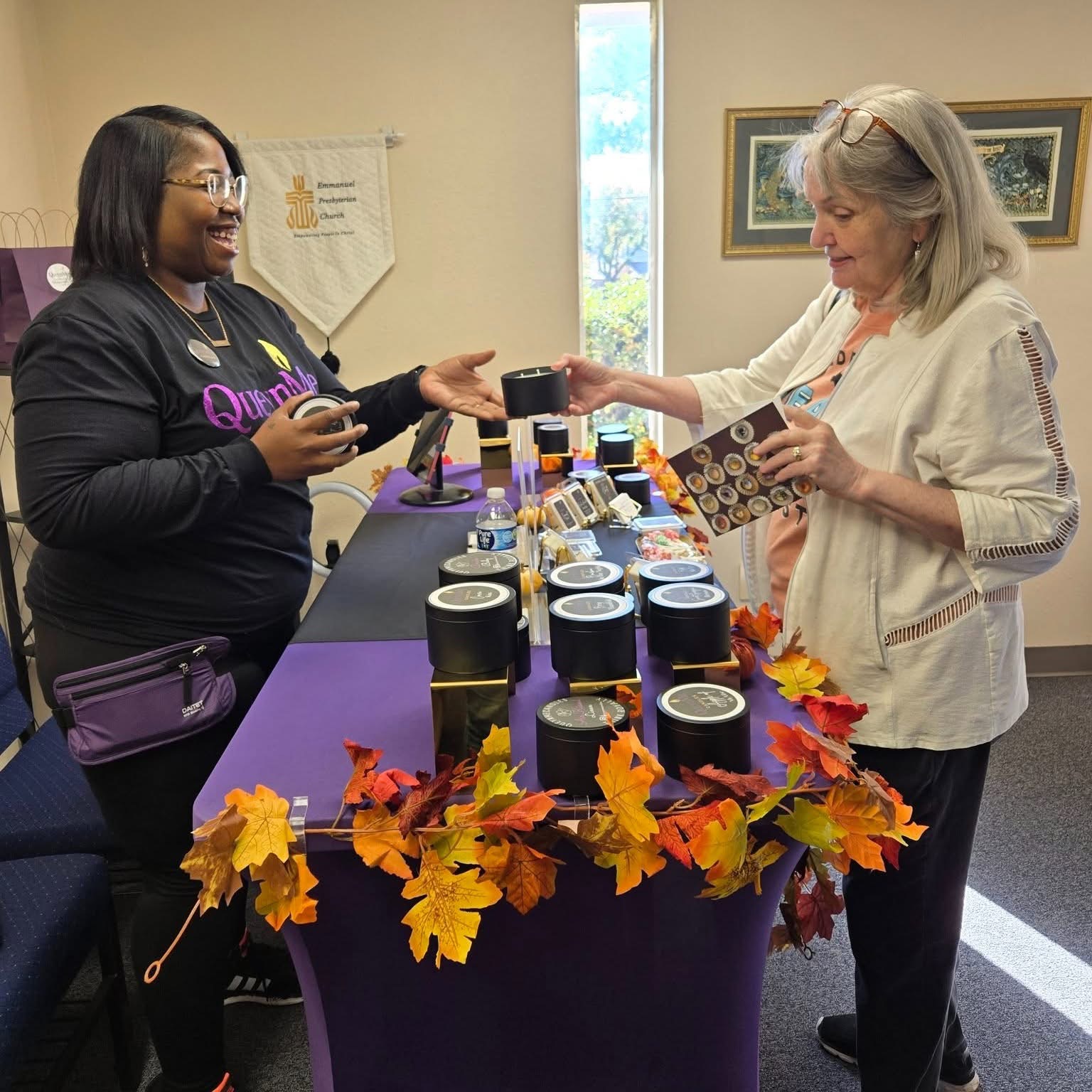 Two women exchanging a cup at a table decorated with autumn leaves, with various items displayed on the table.