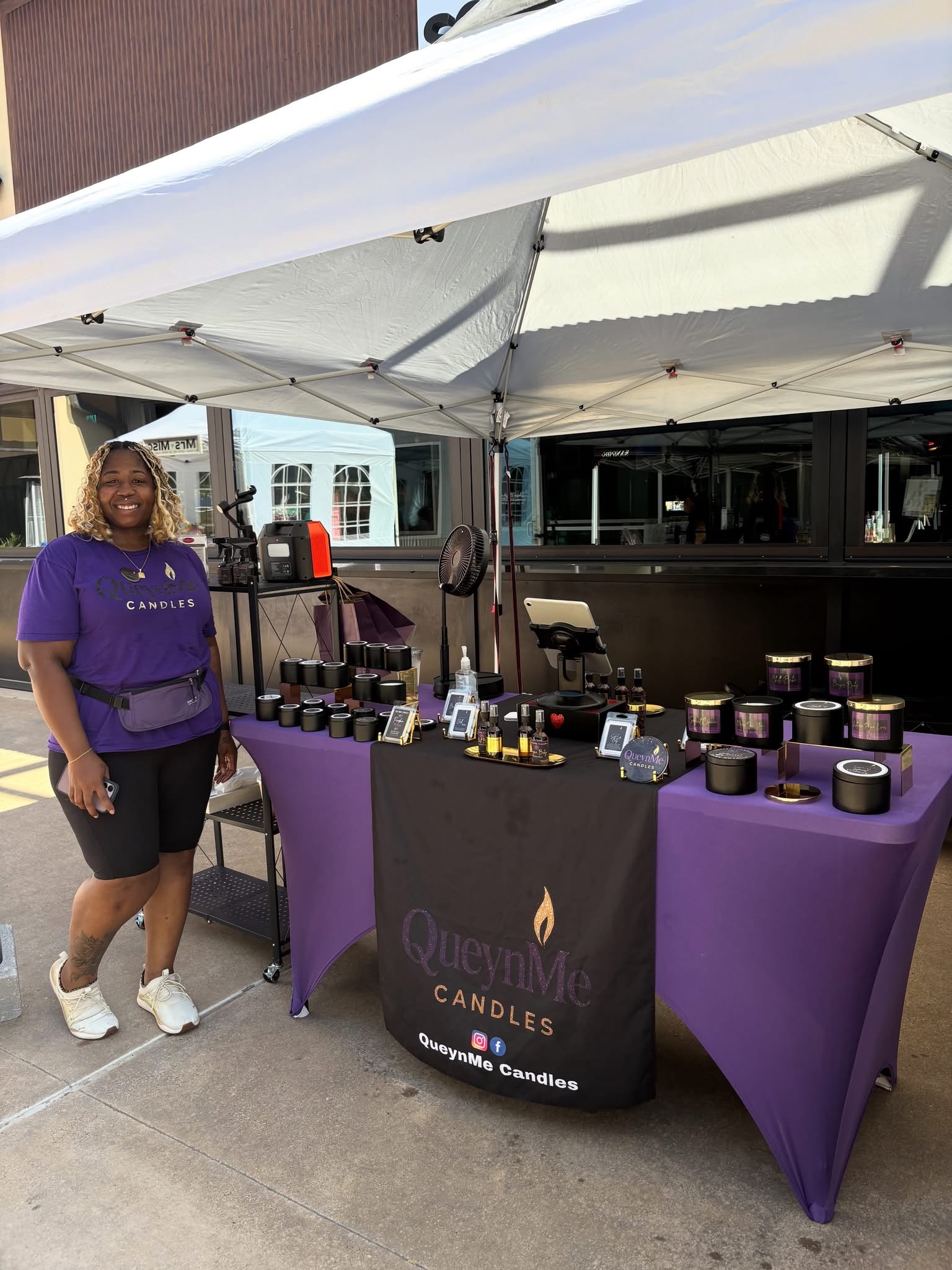 Woman standing next to a table with candles and products under a white canopy outside, building in background.