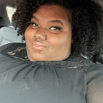 Person with curly hair taking a selfie in a car, smiling slightly, wearing a dark top.