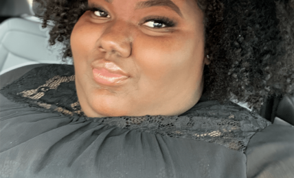 Person with curly hair taking a selfie in a car, smiling slightly, wearing a dark top.