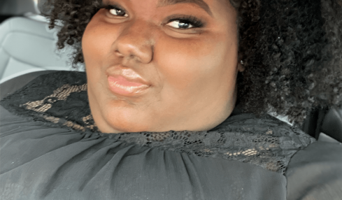 Person with curly hair taking a selfie in a car, smiling slightly, wearing a dark top.