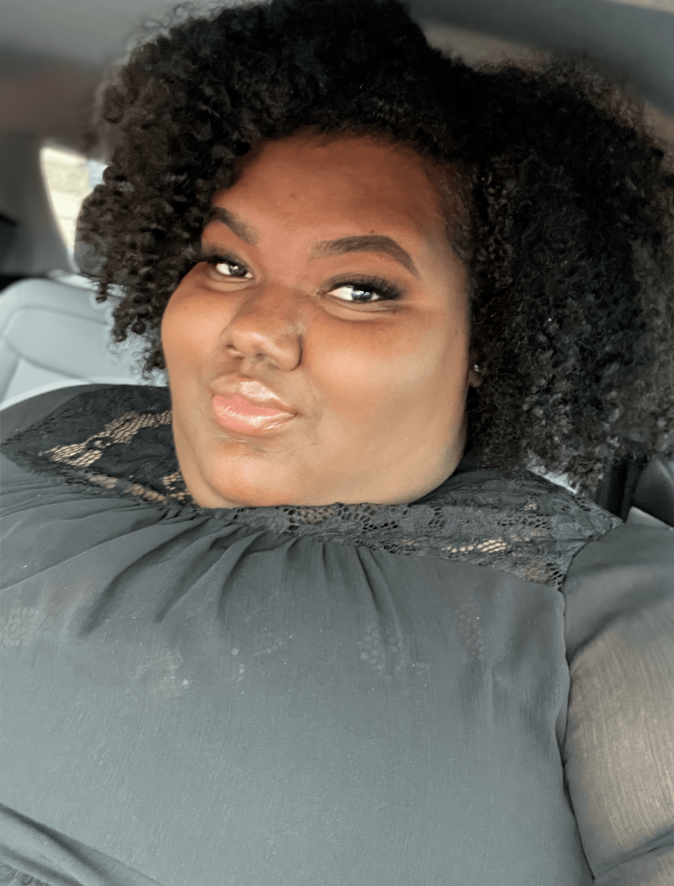 Person with curly hair taking a selfie in a car, smiling slightly, wearing a dark top.