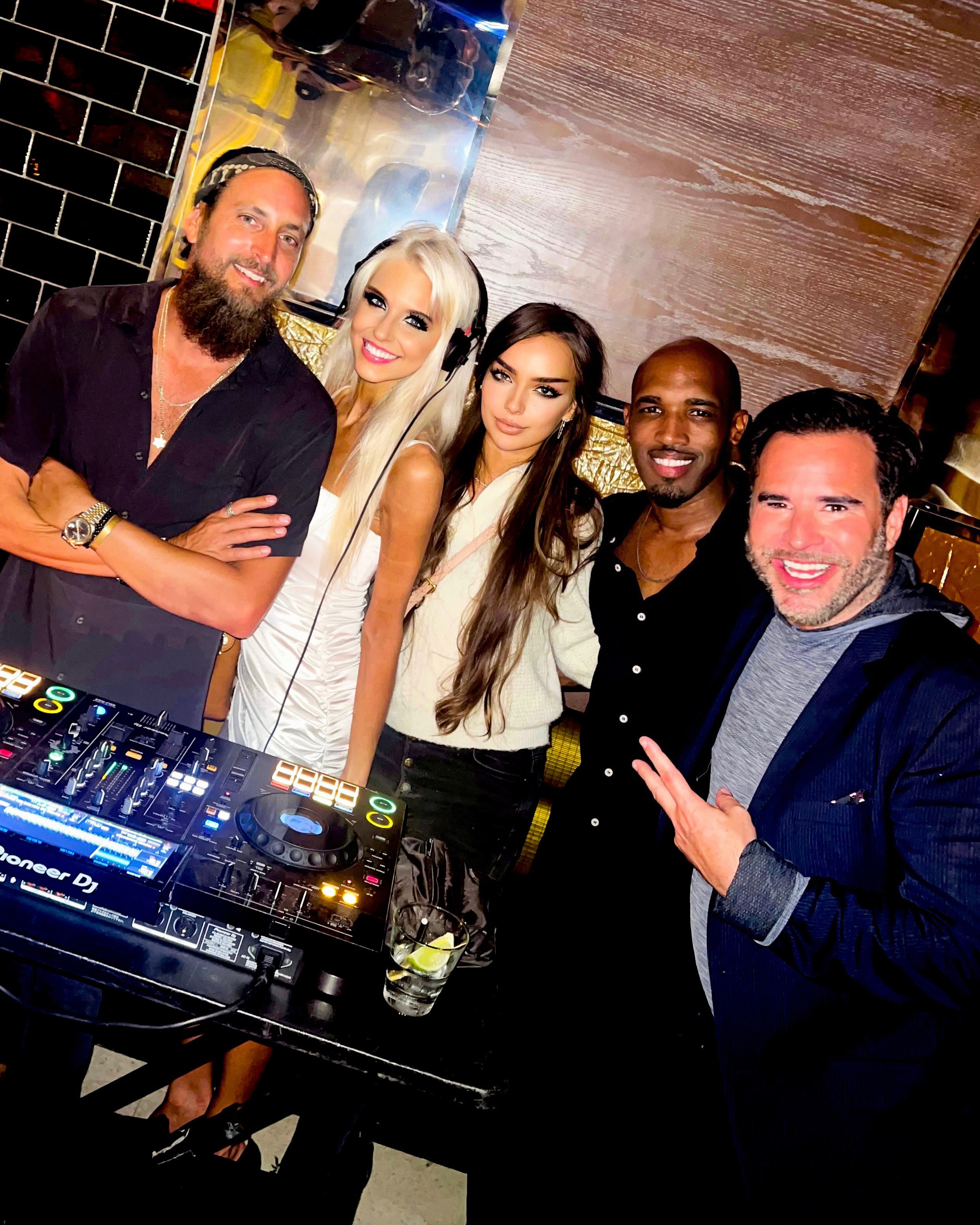 Group of five people smiling and posing behind DJ equipment in a dimly lit indoor setting.