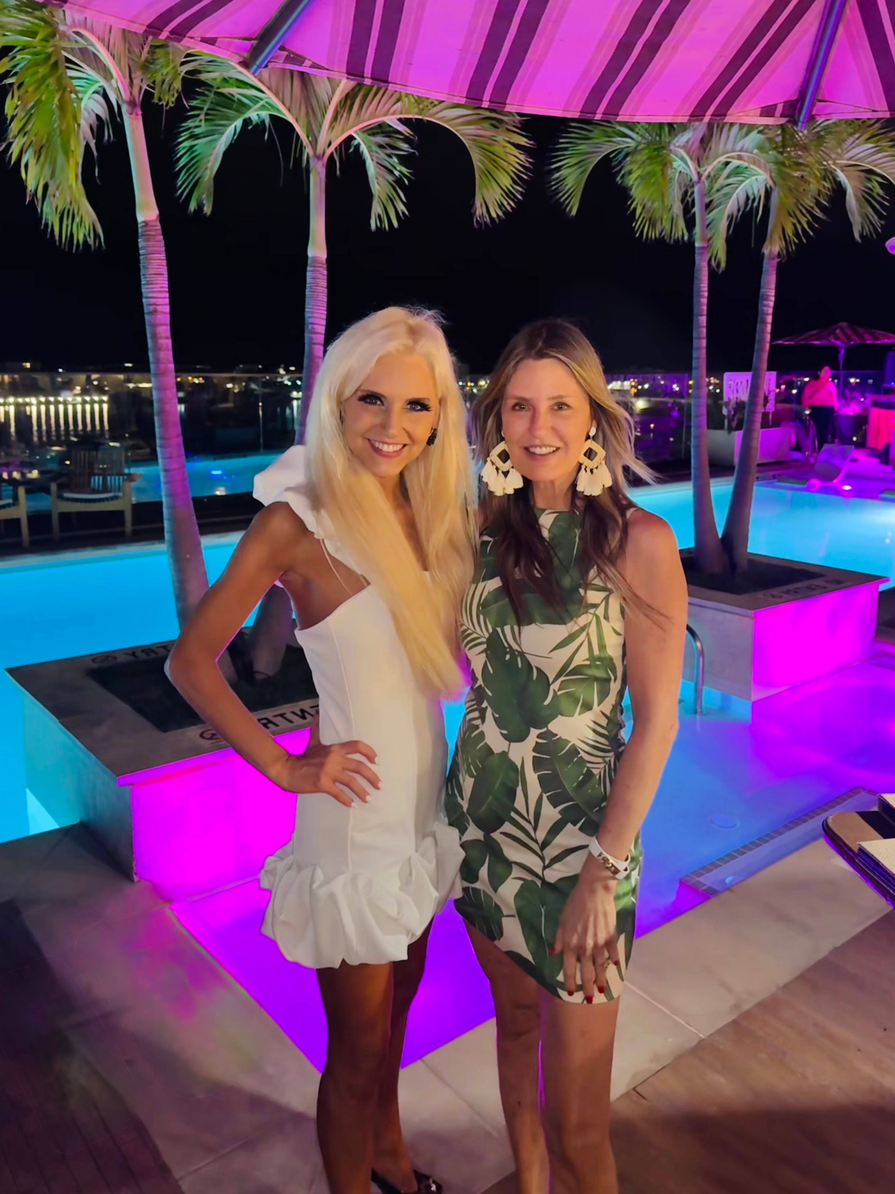 Two women smiling under a pink umbrella with palm trees and a lit pool in the background.