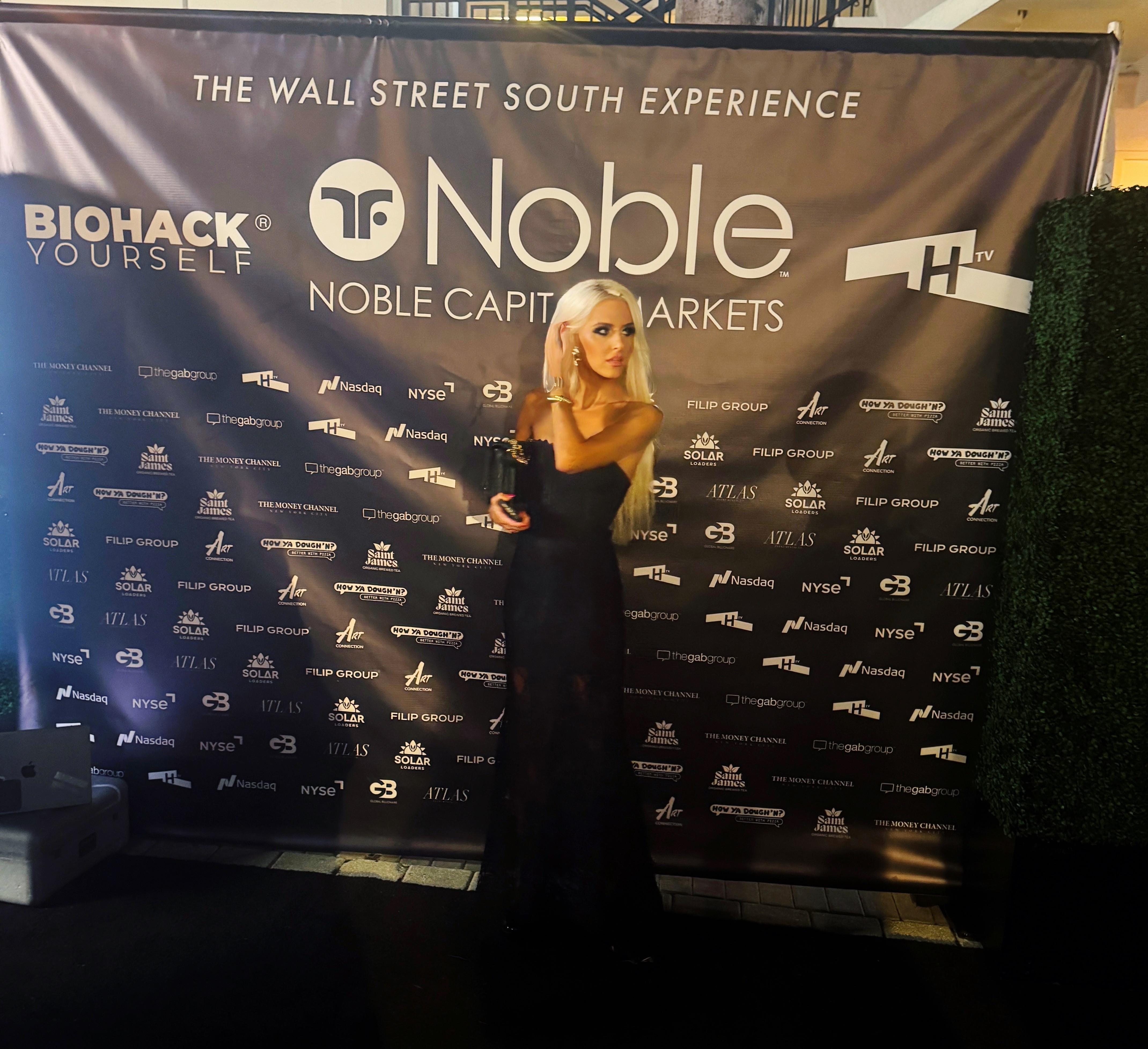 Woman in black dress holding an award stands in front of a backdrop with logos and text.