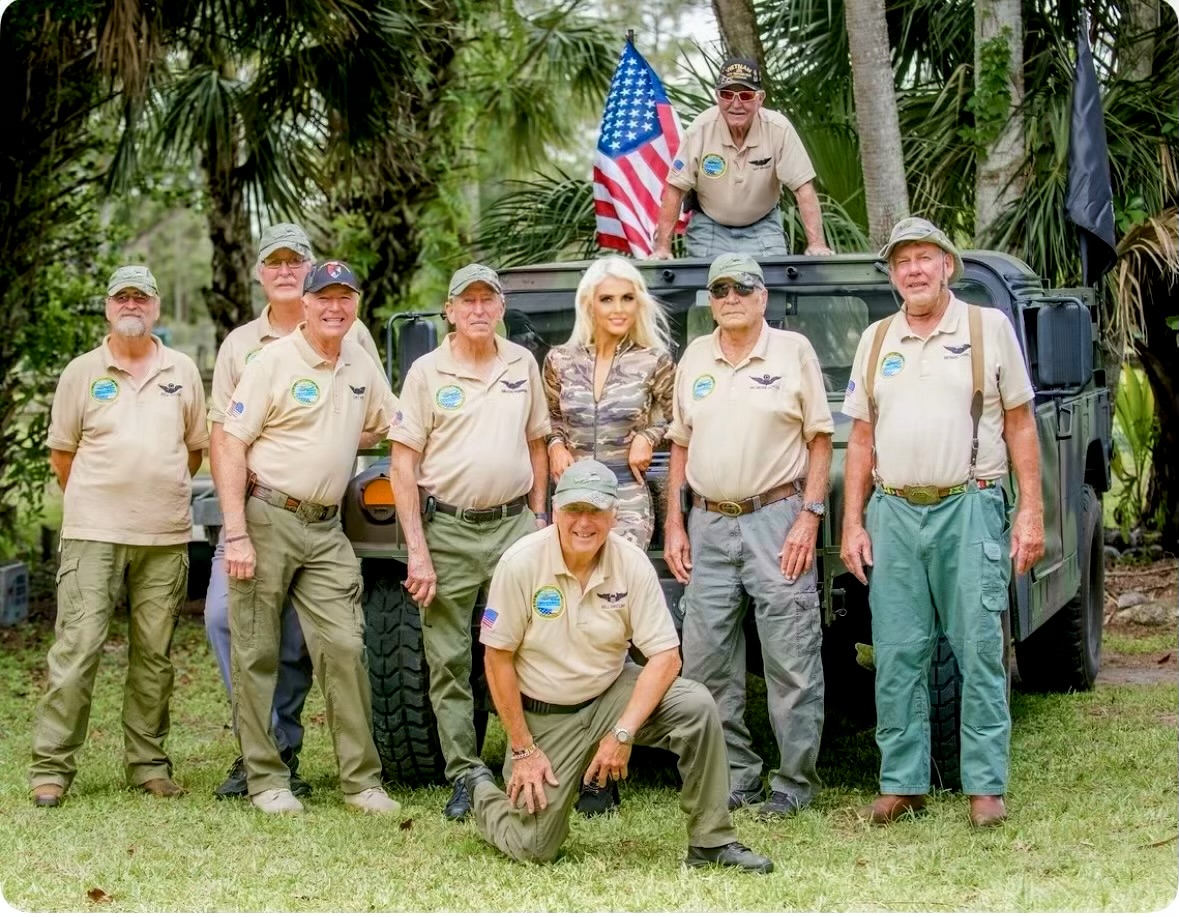 Group of nine people outdoors, some wearing beige shirts, one person sitting on a vehicle, American flag, trees in background.