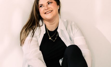 Woman sitting on the floor, smiling, wearing a white shirt, black pants, and white sneakers, against a plain background.