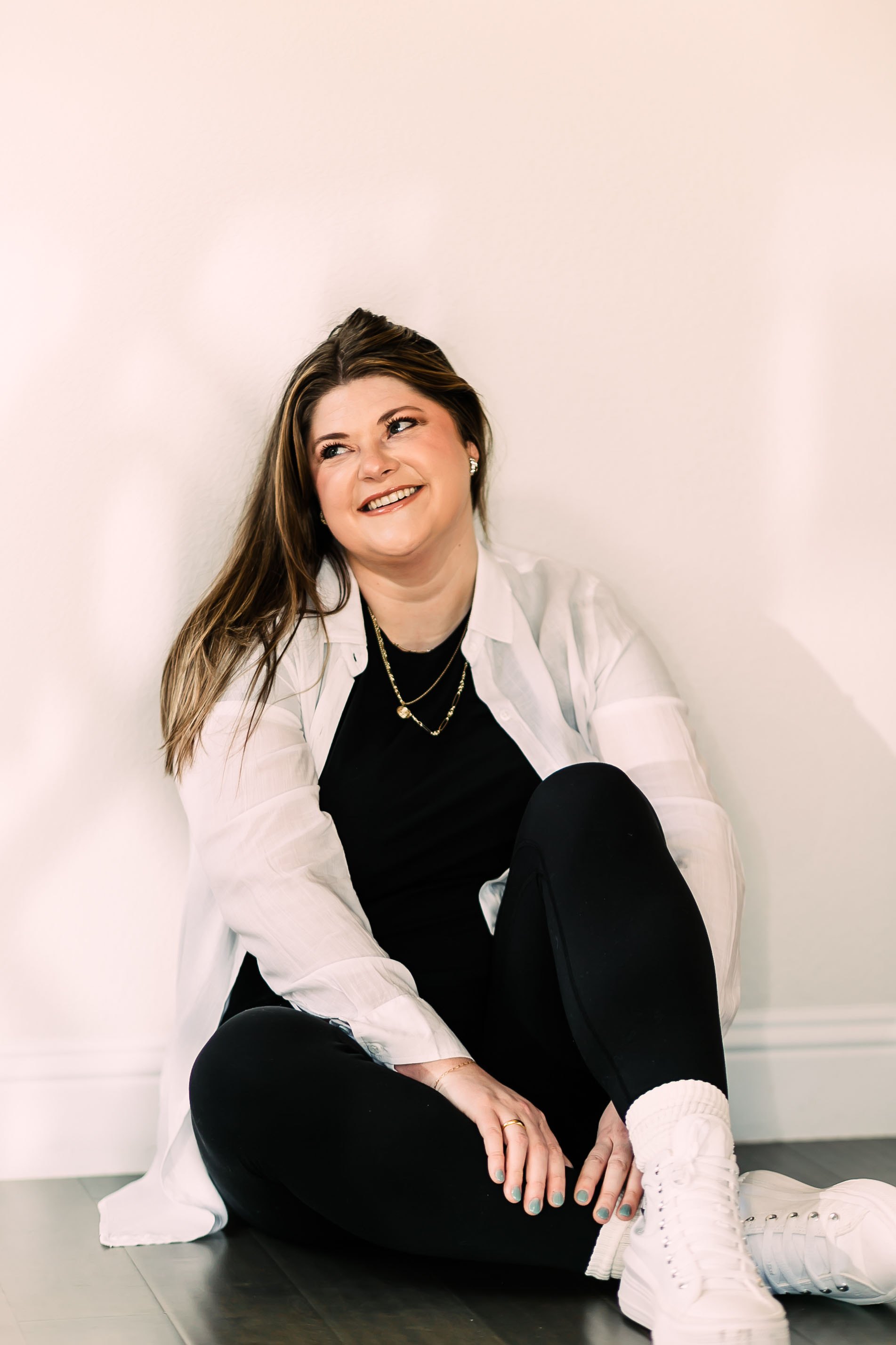 Woman sitting on the floor, smiling, wearing a white shirt, black pants, and white sneakers, against a plain background.