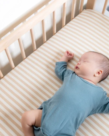 Baby sleeping on a striped mattress in a crib, wearing a blue outfit, with arm raised and eyes closed.
