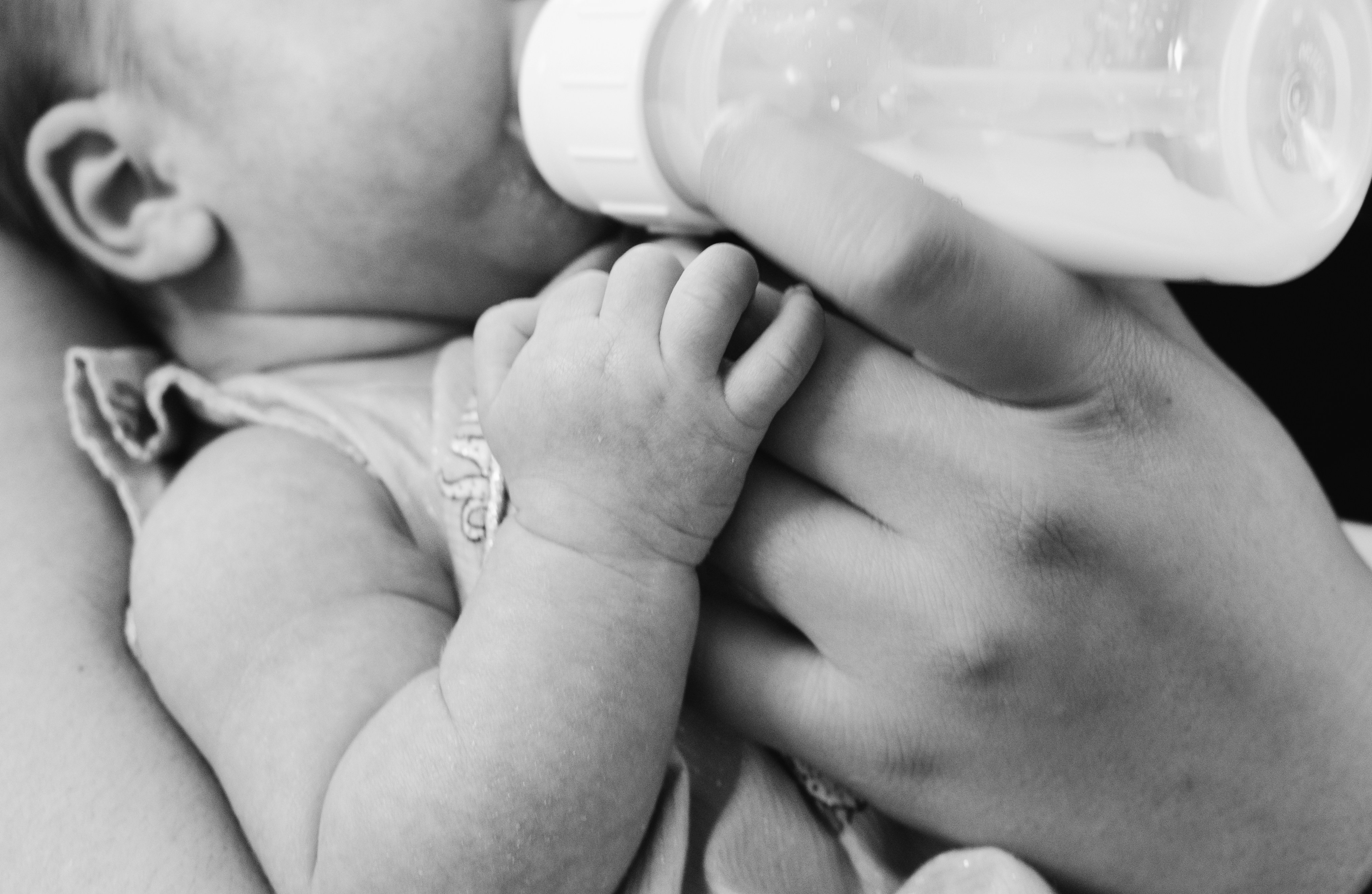 Baby drinking from a bottle held by an adult's hand, close-up in black and white.
