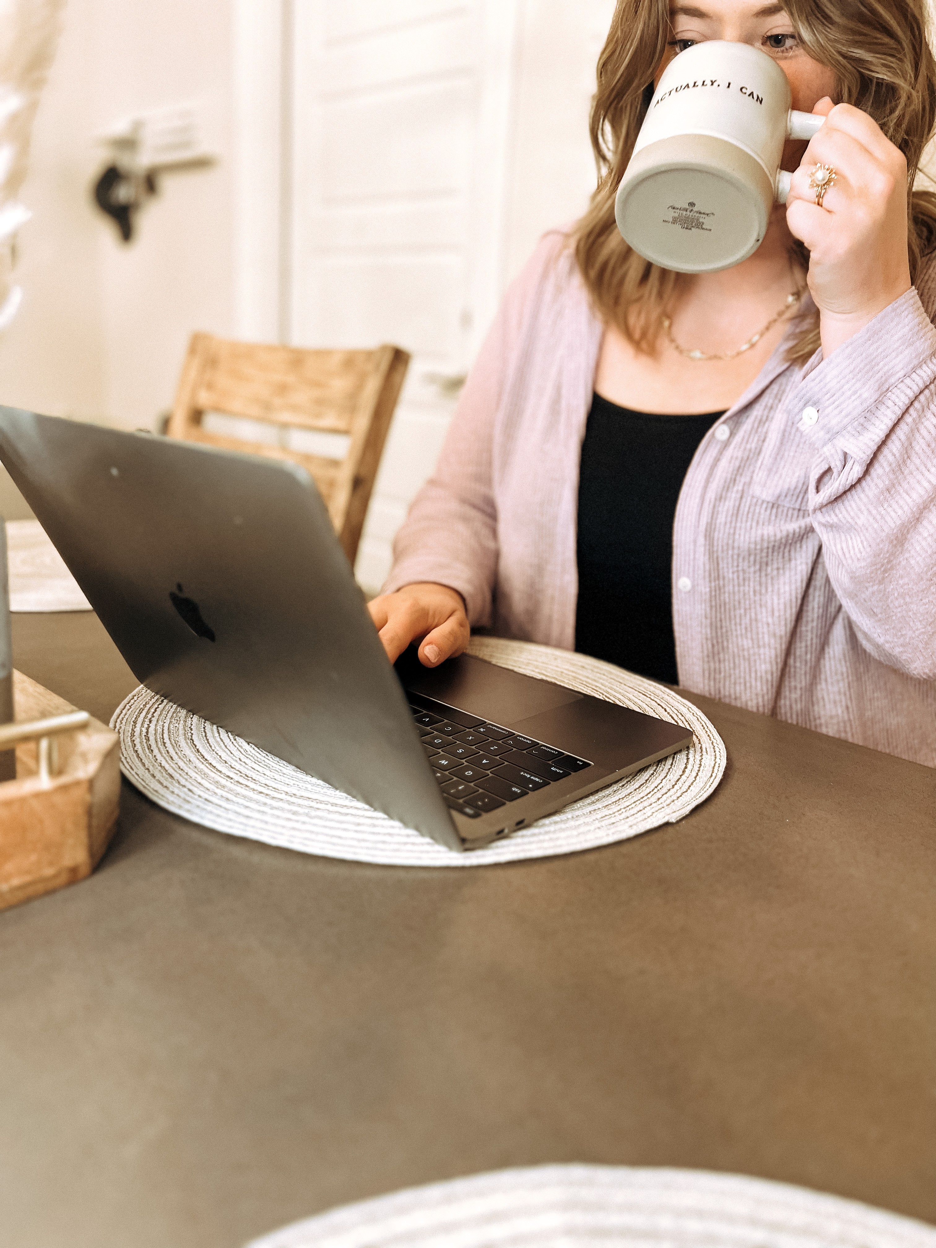 Person drinking coffee while using a laptop at a table in a cozy room.