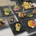 Assorted Japanese dishes including sashimi, tempura, and sushi on black plates, arranged on a white table.