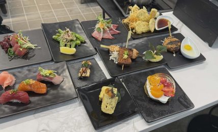 Assorted Japanese dishes including sashimi, tempura, and sushi on black plates, arranged on a white table.