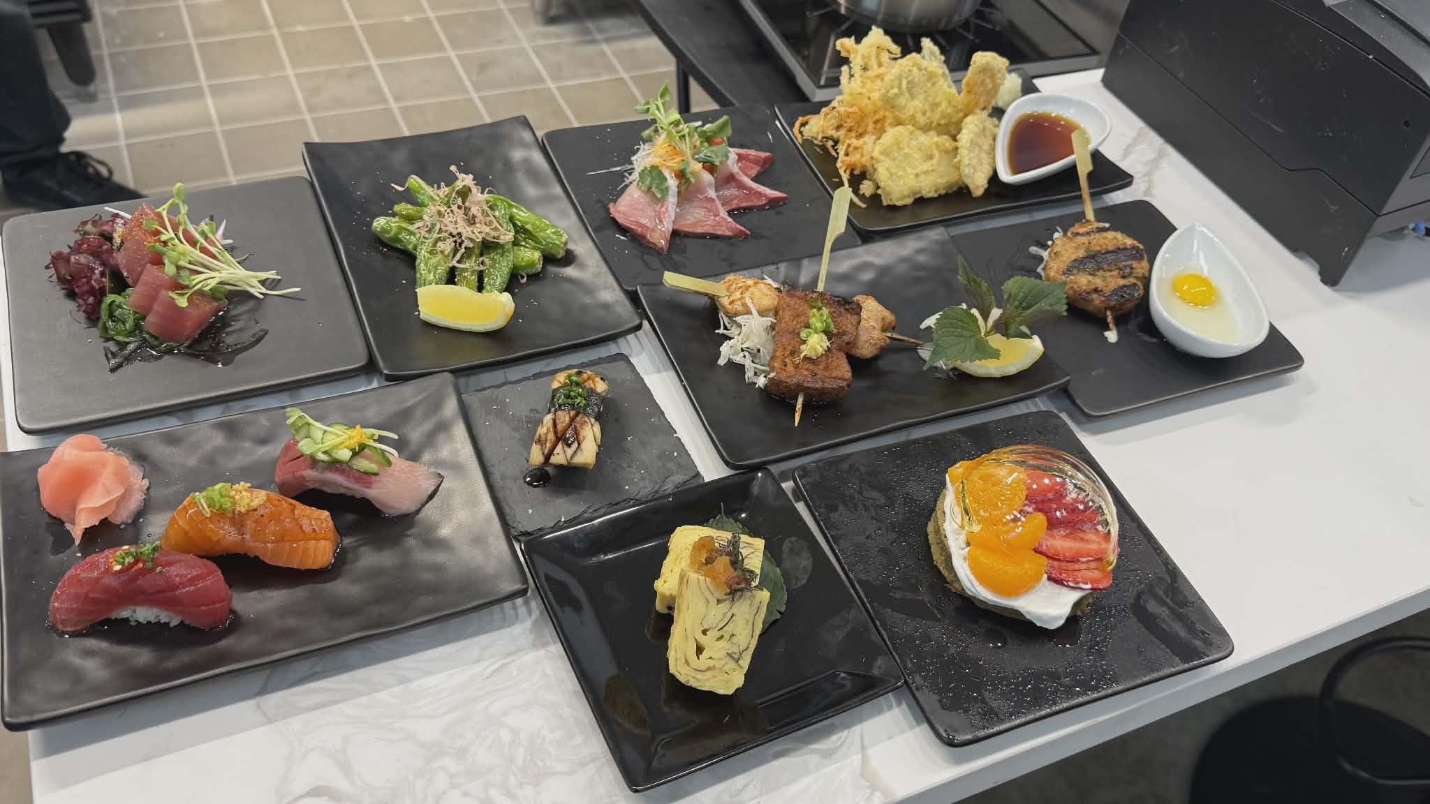 Assorted Japanese dishes including sashimi, tempura, and sushi on black plates, arranged on a white table.