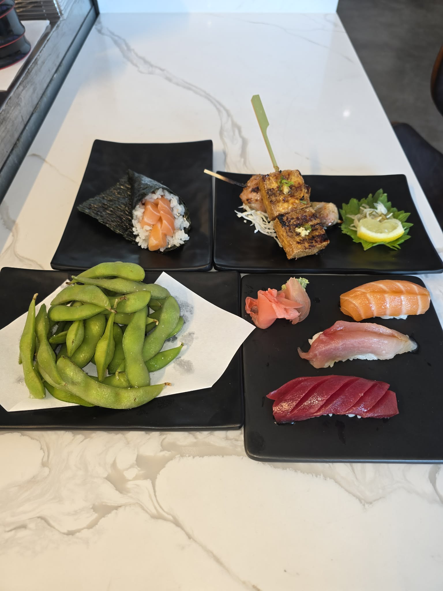 Assorted sushi and edamame on black plates, with a sushi roll, sashimi, and garnishes on a white marble surface.