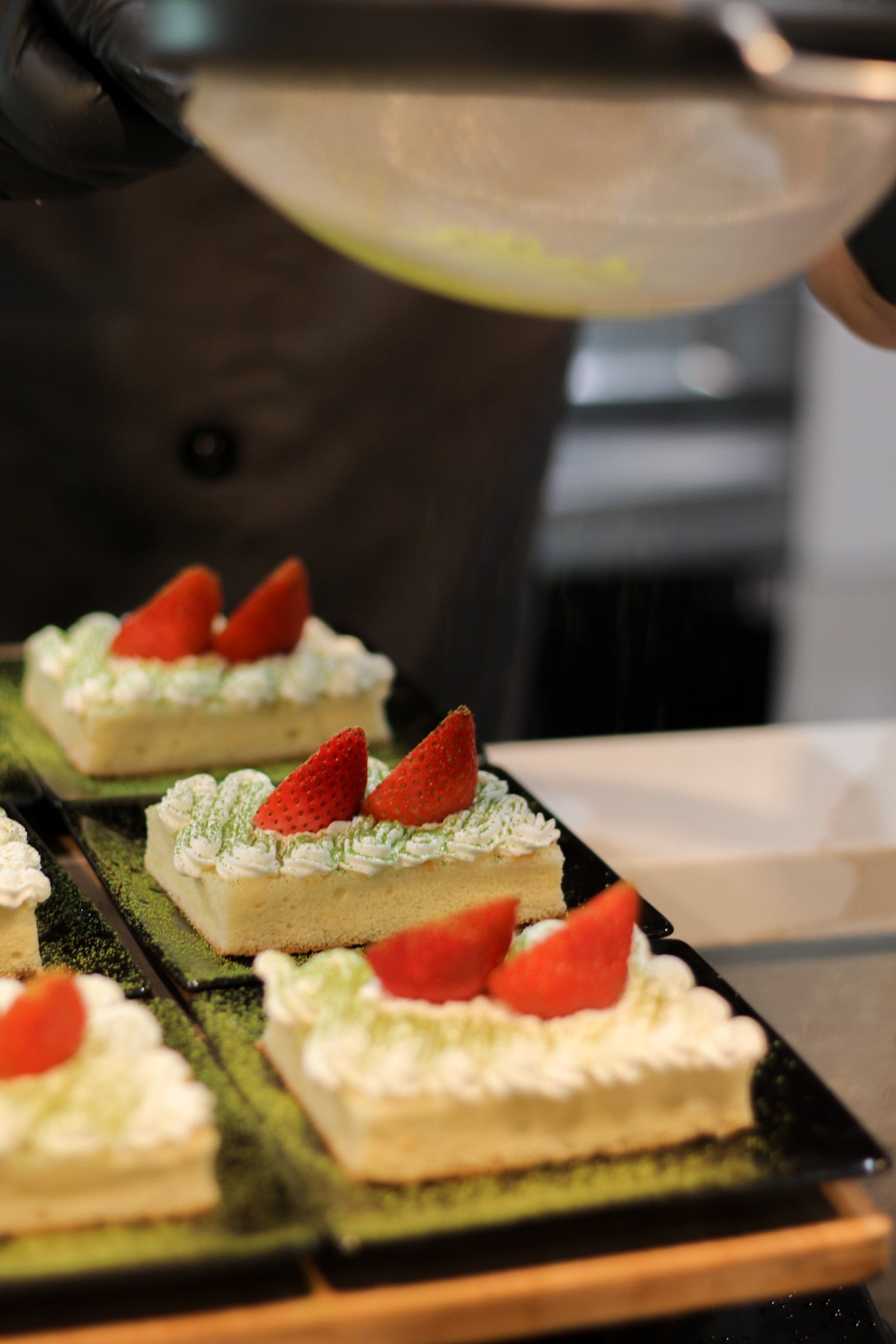 Four rectangular desserts topped with whipped cream and strawberries on a black tray, with a person pouring liquid in the background.