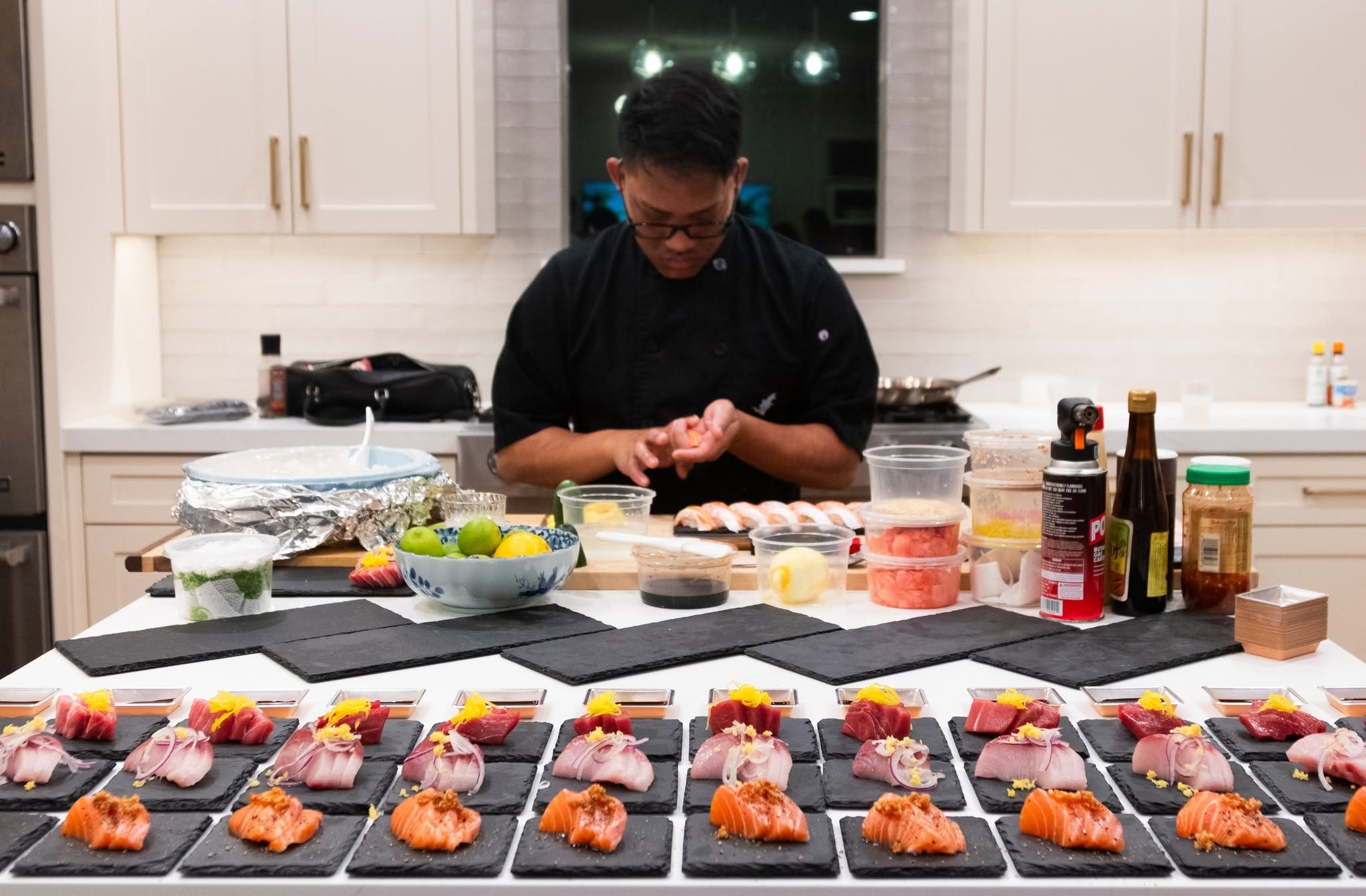 Person preparing food in a kitchen with multiple plates of sushi in front, various ingredients and utensils on the counter.