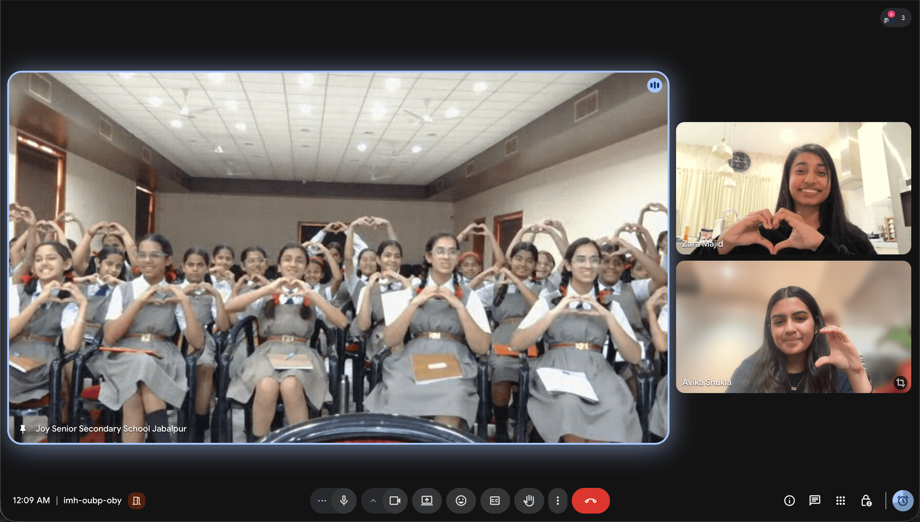 Group of students in classroom, some making heart shapes with hands, seated at desks, with a teacher or presenter at the front.