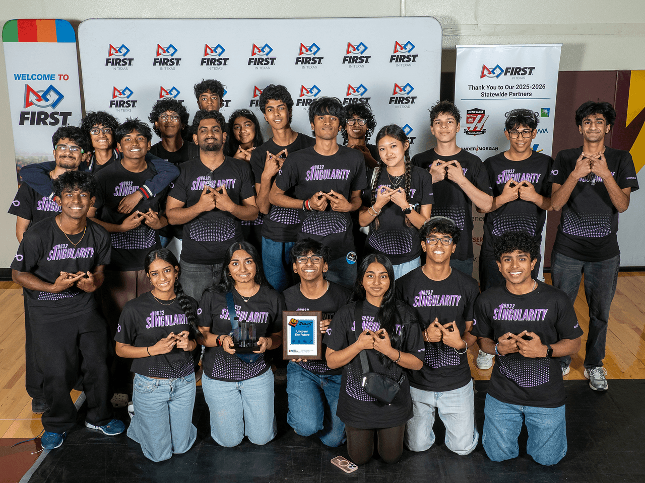Group of young people in matching shirts holding awards, standing in front of banners at an event.