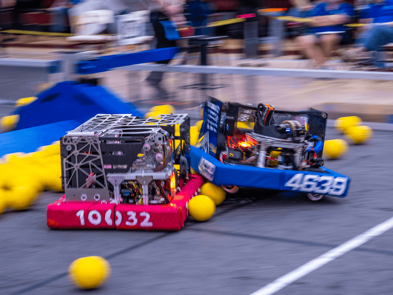 Two robots compete in a robotics competition, with yellow balls scattered on the floor and spectators in the background.