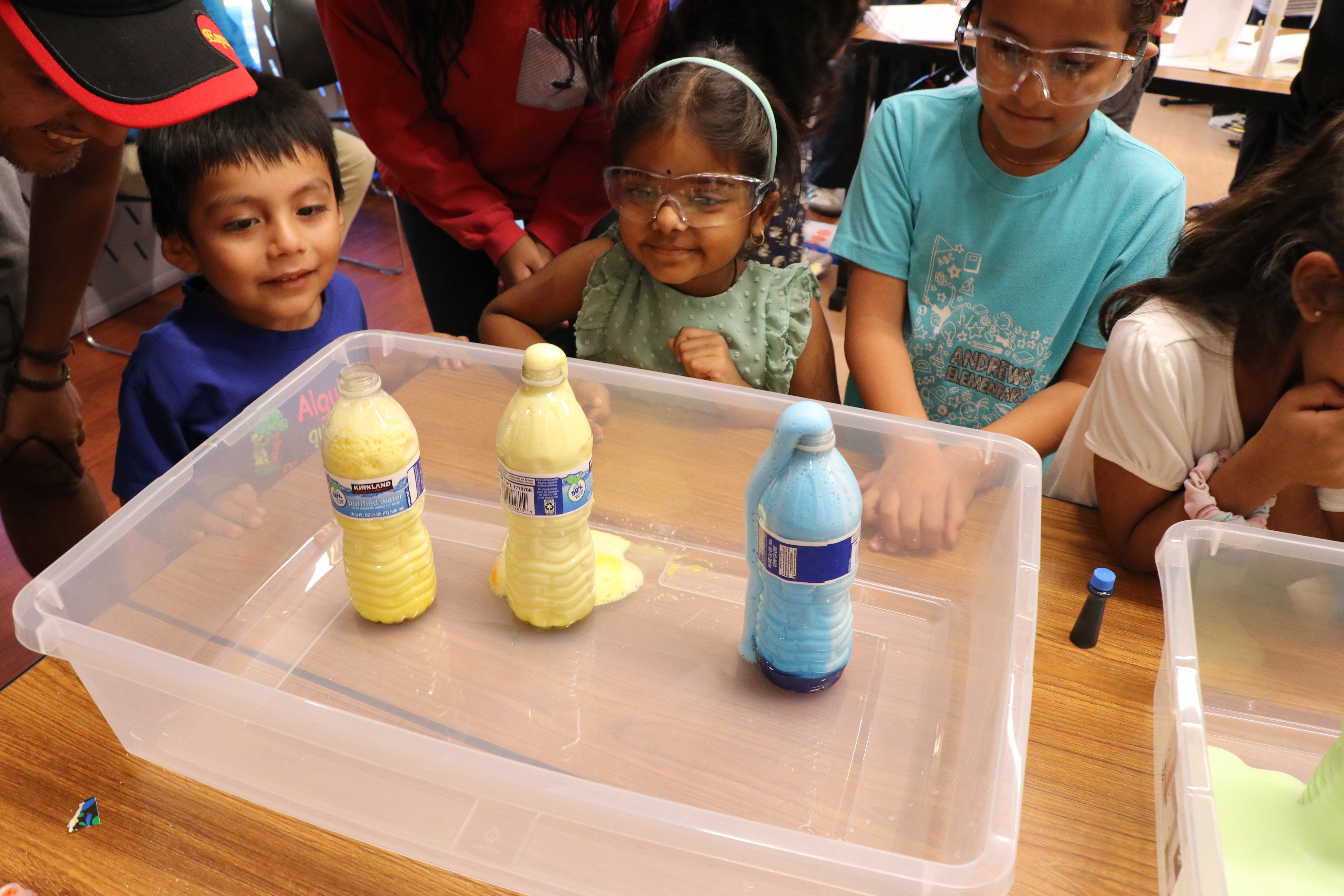 Children observe bottles with yellow and blue labels inside a clear plastic container, with some adults nearby.