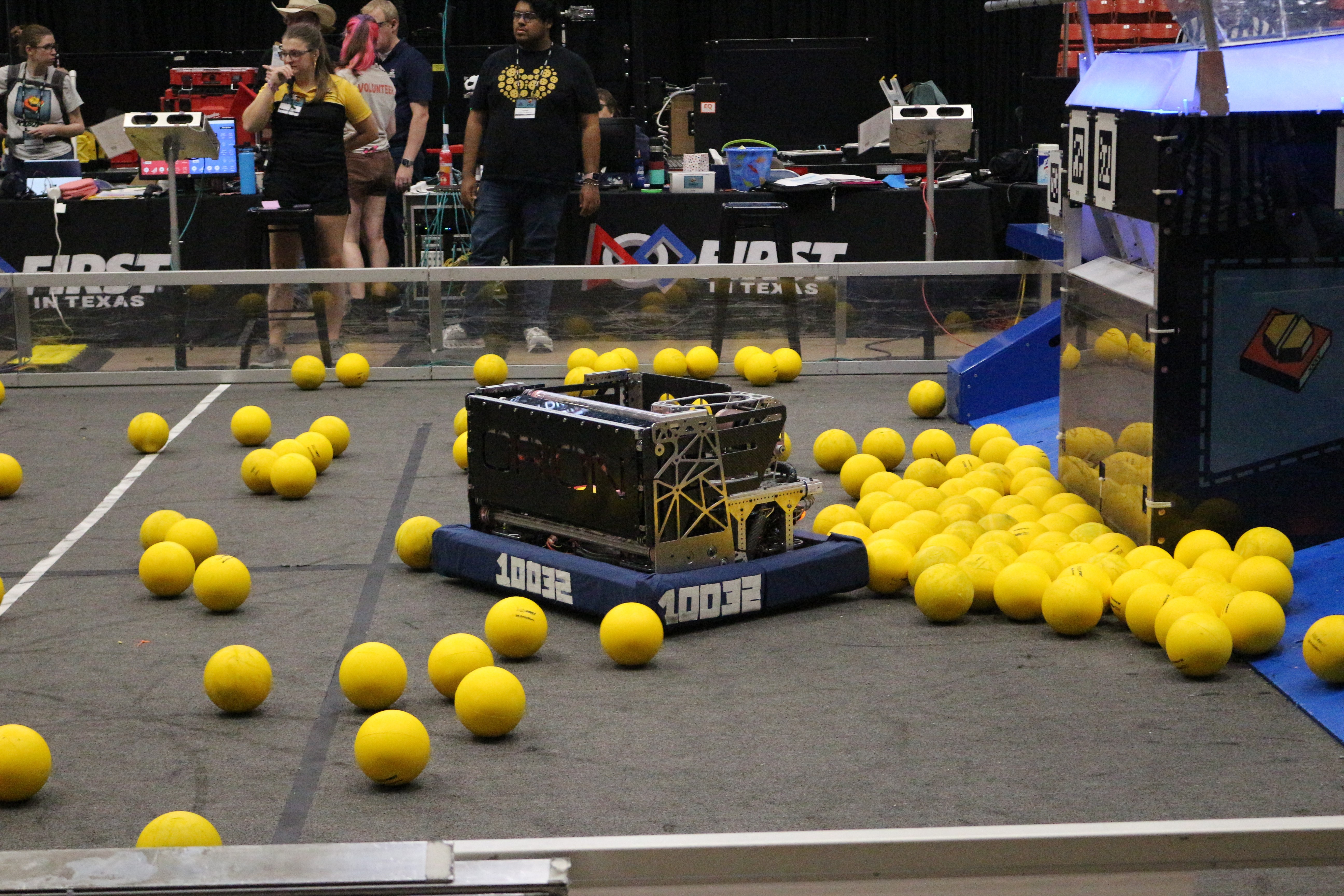 Robotic competition field with yellow balls scattered, a robot in the center, and people observing in the background.
