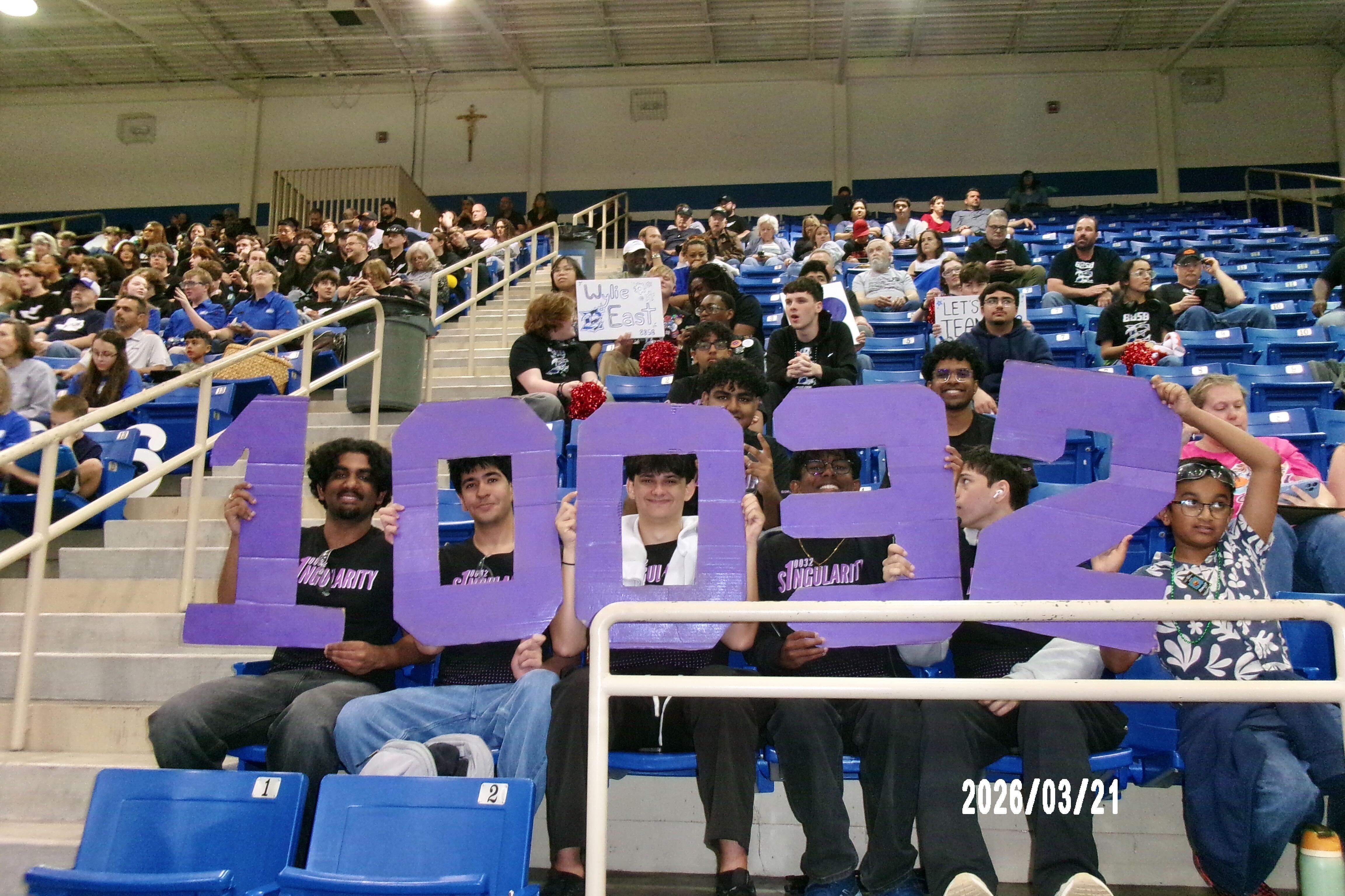 Group of people holding large purple numbers '100' in a gymnasium with blue seats and a cross on the wall, March 21, 2023.