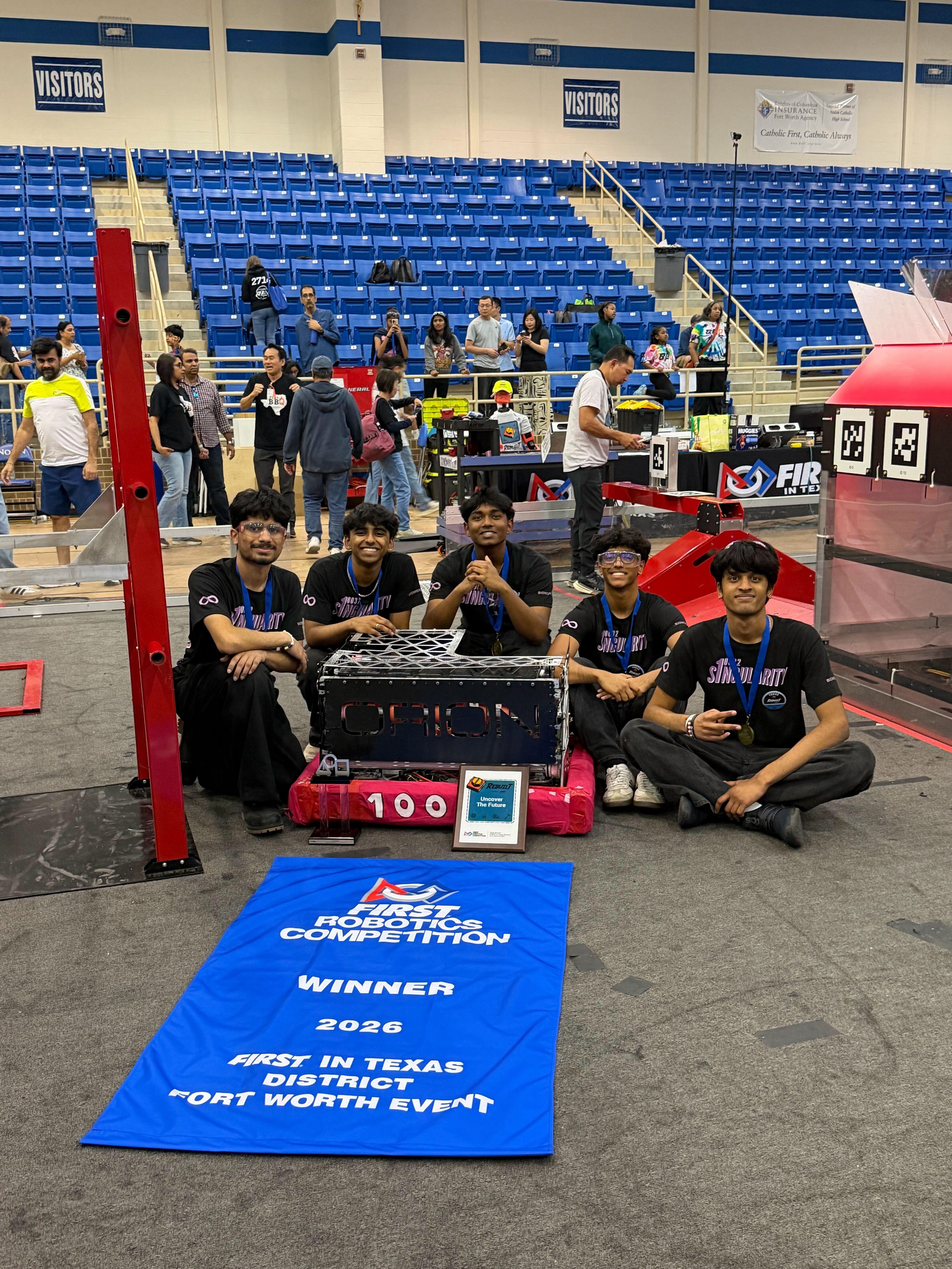 Group of five people kneeling with a robot and trophy at a robotics competition, blue winner mat in front, indoor arena setting.