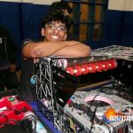 Person smiling with arms resting on a DJ equipment setup, another person in the background, indoor setting, date visible.