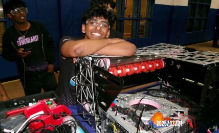 Person smiling with arms resting on a DJ equipment setup, another person in the background, indoor setting, date visible.