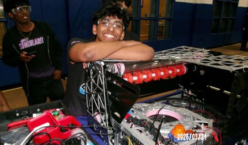 Person smiling with arms resting on a DJ equipment setup, another person in the background, indoor setting, date visible.