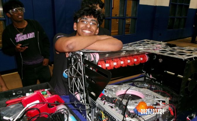Person smiling with arms resting on a DJ equipment setup, another person in the background, indoor setting, date visible.