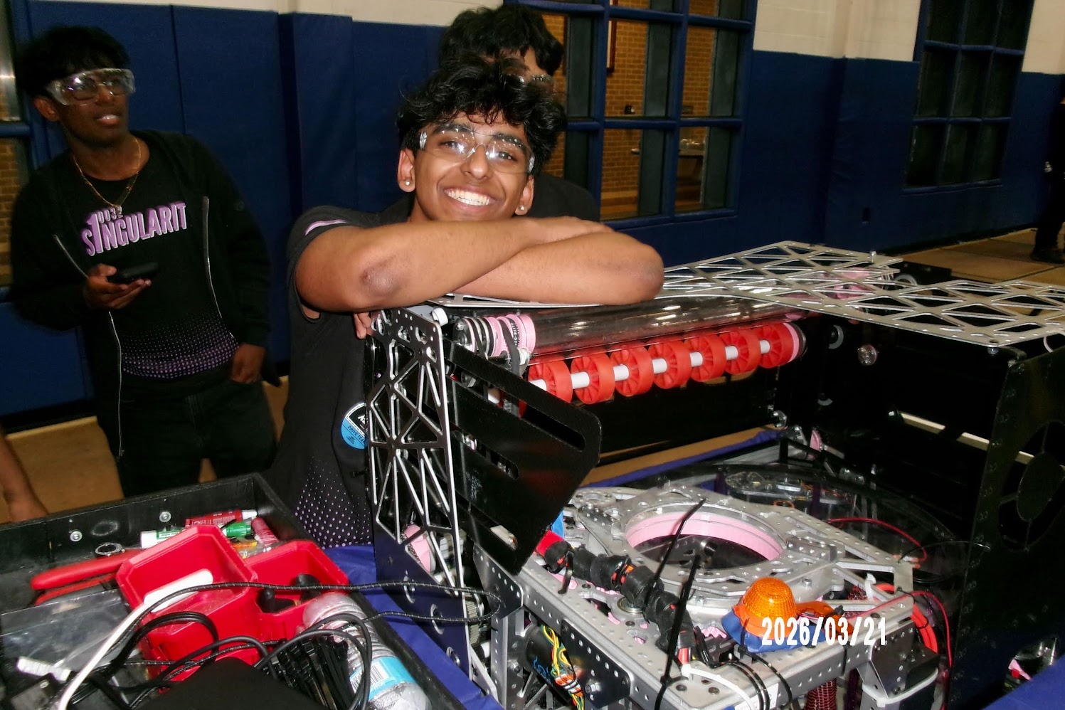 Person smiling with arms resting on a DJ equipment setup, another person in the background, indoor setting, date visible.