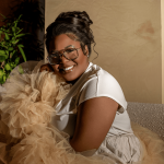 Woman smiling, wearing glasses, sitting on a sofa with a fluffy dog, in a cozy indoor setting.